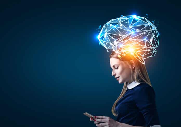 Side view of a blond businesswoman looking at her smartphone screen standing near a dark blue wall. There is a large blue brain hologram around her head.